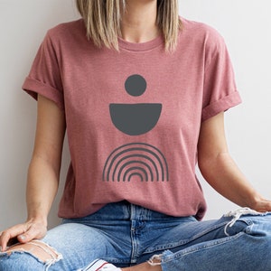 May include: A dusty rose t-shirt with rolled sleeves. The shirt features a dark grey abstract design of a circle, a half-circle, and a rainbow. The person is wearing blue jeans.