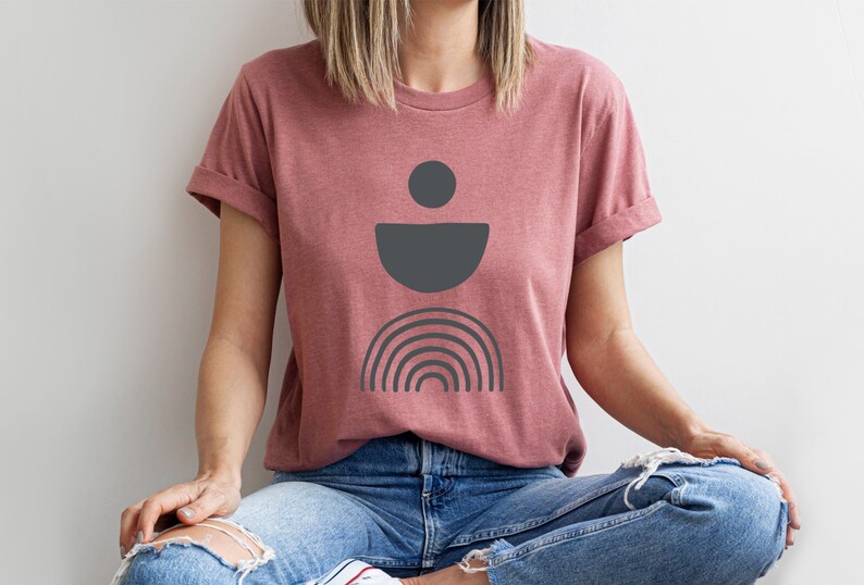 May include: A dusty rose t-shirt with rolled sleeves. The shirt features a dark grey abstract design of a circle, a half-circle, and a rainbow. The person is wearing blue jeans.