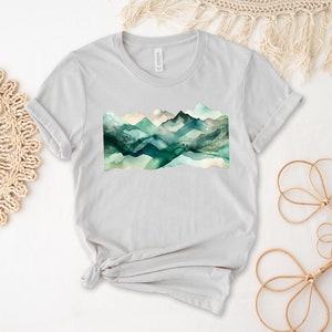 Puede incluir: Una camiseta gris claro con un dise&ntilde;o de acuarela de monta&ntilde;as verdes y azules.