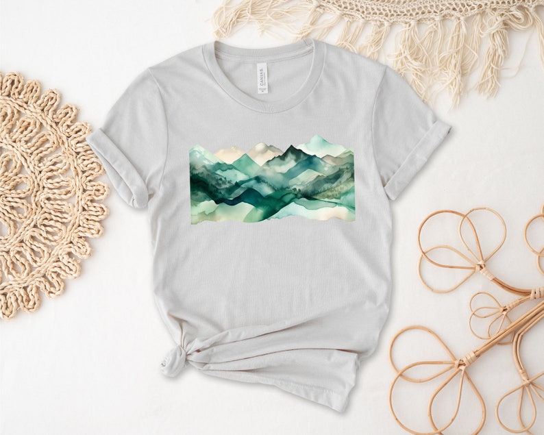 Puede incluir: Una camiseta gris claro con un dise&ntilde;o de acuarela de monta&ntilde;as verdes y azules.