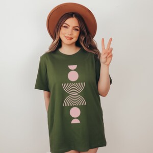 May include: A green t-shirt with a pink and brown geometric design. The design features a series of circles, half-circles, and arcs stacked vertically.