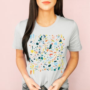 Pu&ograve; includere: Una t-shirt bianca con un design a motivi di terrazzo colorati. Il motivo presenta diverse forme e colori, tra cui blu, verde, giallo, arancione e rosa.