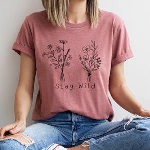 May include: A mauve t-shirt with a black line drawing of two bouquets of flowers and the text "Stay Wild".
