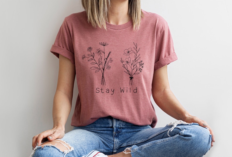 May include: A mauve t-shirt with a black line drawing of two bouquets of flowers and the text "Stay Wild".