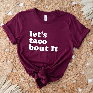 May include: Burgundy t-shirt with white text that reads "let's taco bout it".