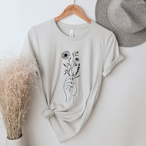 May include: A light gray t-shirt with a black line drawing of a hand holding a bouquet of flowers.