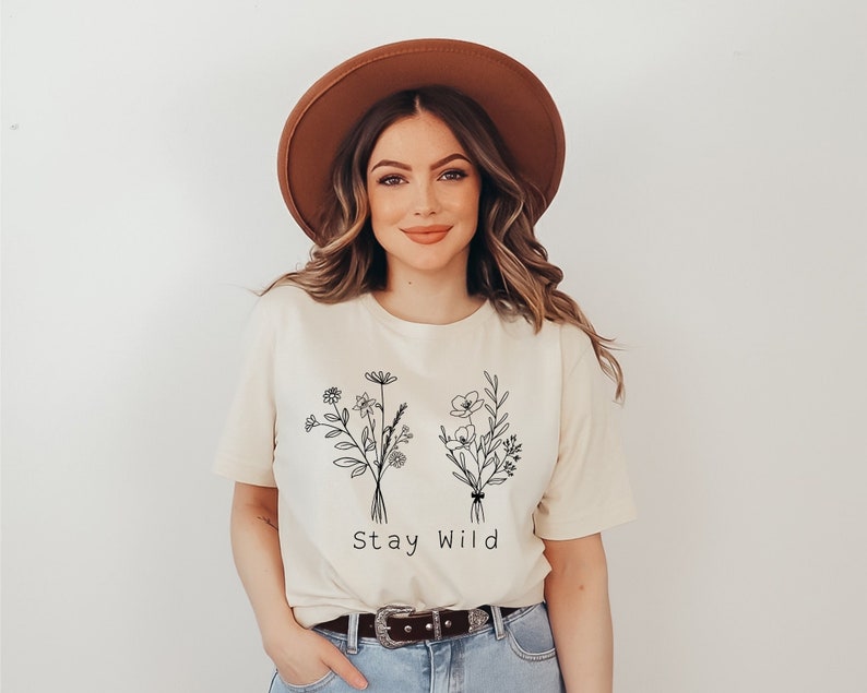 A short sleeve t-shirt with a minimalist floral graphic of two wildflower bouquets. Under it says "Stay Wild". Entire design is in black. Shirt is the Bella Canvas 3001 unisex jersey tee. Very high quality and soft.