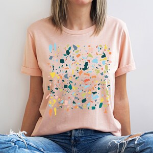 Pu&ograve; includere: Una maglietta rosa con un design a stampa di terrazzo colorato. La stampa presenta varie forme e colori, tra cui blu, verde, giallo, arancione e rosa.