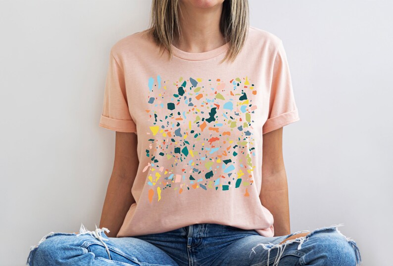 Pu&ograve; includere: Una maglietta rosa con un design a stampa di terrazzo colorato. La stampa presenta varie forme e colori, tra cui blu, verde, giallo, arancione e rosa.