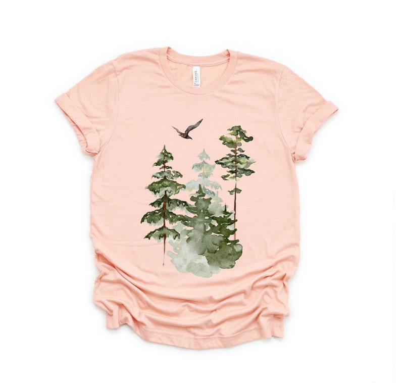 May include: A peach-colored t-shirt with a watercolor design of a bird flying over a forest. The trees are green and have a misty effect.