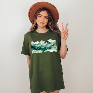 Puede incluir: Una camiseta verde con un dise&ntilde;o de cordillera de monta&ntilde;as de acuarela. Las monta&ntilde;as son de tonos azules, verdes y blancos. La camiseta tiene un corte holgado y cuello redondo.