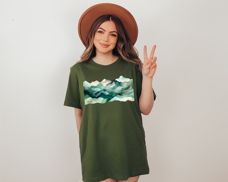 Puede incluir: Una camiseta verde con un dise&ntilde;o de cordillera de monta&ntilde;as de acuarela. Las monta&ntilde;as son de tonos azules, verdes y blancos. La camiseta tiene un corte holgado y cuello redondo.