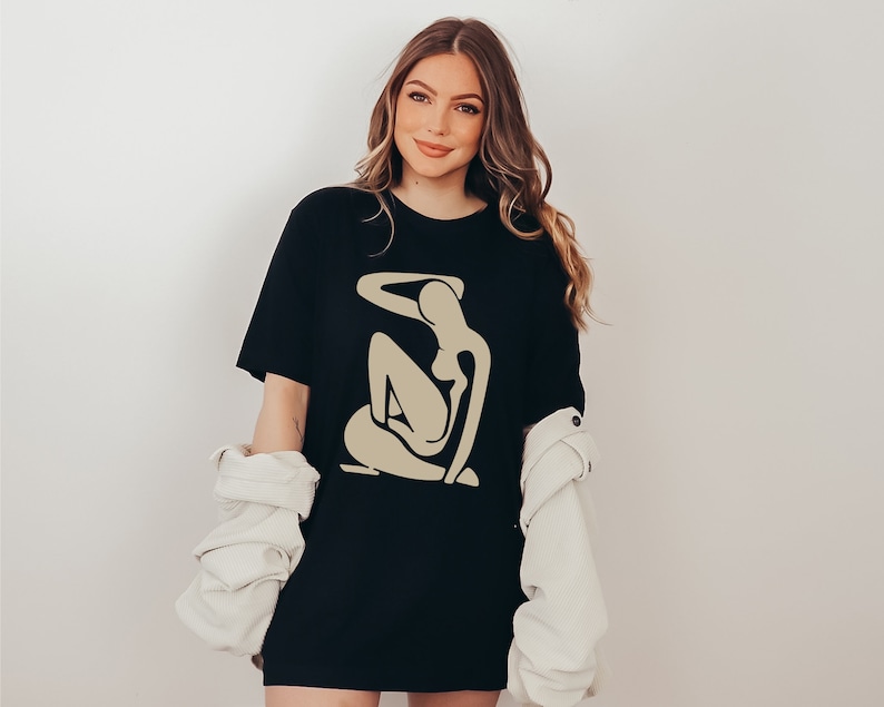 May include: A black t-shirt with a beige abstract figure design. The figure is a stylized depiction of a person sitting with their arm raised above their head.