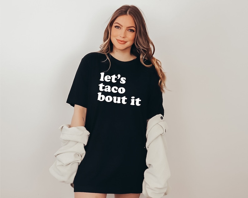 May include: A black t-shirt with white text that reads "let's taco bout it" in a retro font.