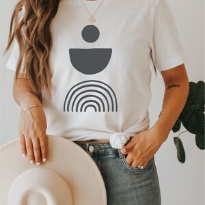 May include: White t-shirt with a dark grey geometric design. The design includes a circle, a half-circle, and a rainbow. The model is wearing blue jeans and holding a cream-coloured hat.