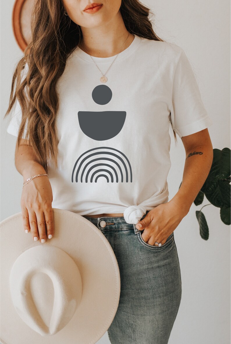 May include: White t-shirt with a dark grey geometric design. The design includes a circle, a half-circle, and a rainbow. The model is wearing blue jeans and holding a cream-coloured hat.