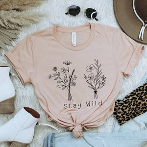 May include: A light pink t-shirt with a black line drawing of two bouquets of flowers and the text "Stay Wild". The shirt is laid out on a white fuzzy surface with a pair of white ankle boots, a brown cowboy hat, a pair of sunglasses, a pair of blue jeans, and a leopard print scarf.