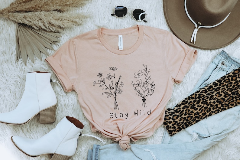 May include: A light pink t-shirt with a black line drawing of two bouquets of flowers and the text "Stay Wild". The shirt is laid out on a white fuzzy surface with a pair of white ankle boots, a brown cowboy hat, a pair of sunglasses, a pair of blue jeans, and a leopard print scarf.