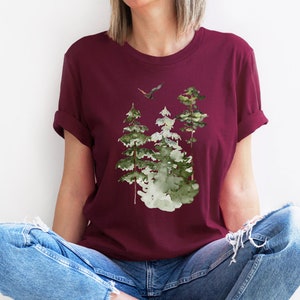 May include: A maroon t-shirt with a watercolor design of three green trees and a bird flying above them.