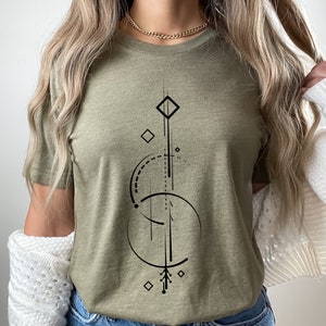 Pode incluir: Uma camiseta verde com um design geom&eacute;trico em preto. O design apresenta uma linha vertical com uma forma de diamante na parte superior, um c&iacute;rculo com uma linha tracejada e uma forma de diamante menor na parte inferior.