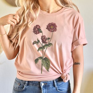May include: A blush pink t-shirt with a floral print of three burgundy flowers with green leaves.