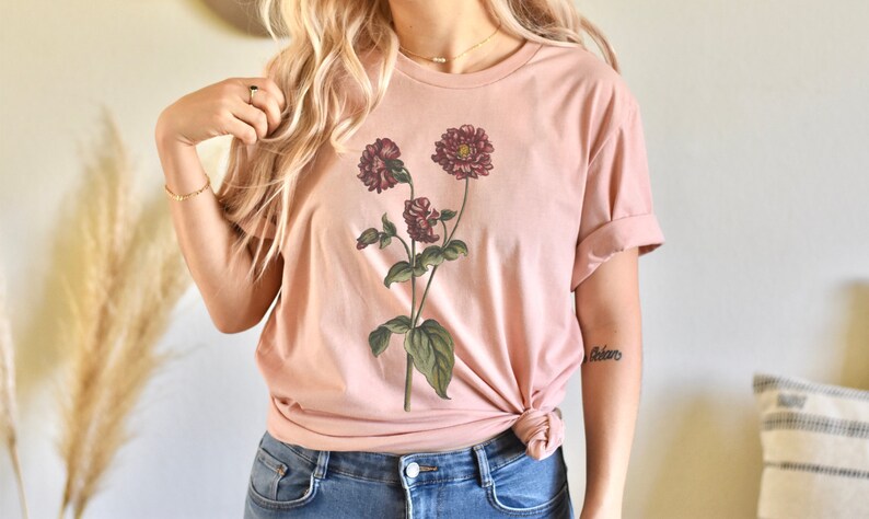 May include: A blush pink t-shirt with a floral print of three burgundy flowers with green leaves.