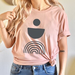 May include: A pale pink t-shirt with a dark grey abstract design. The design features a circle, a half-circle, and a rainbow. The shirt is knotted at the waist and worn with blue jeans.