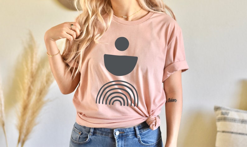May include: A pale pink t-shirt with a dark grey abstract design. The design features a circle, a half-circle, and a rainbow. The shirt is knotted at the waist and worn with blue jeans.