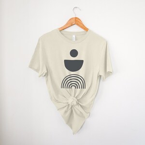 May include: A cream-coloured t-shirt with a dark grey geometric design featuring a circle, a half-circle, and a rainbow. The t-shirt is knotted at the waist and hangs on a wooden hanger against a white background.