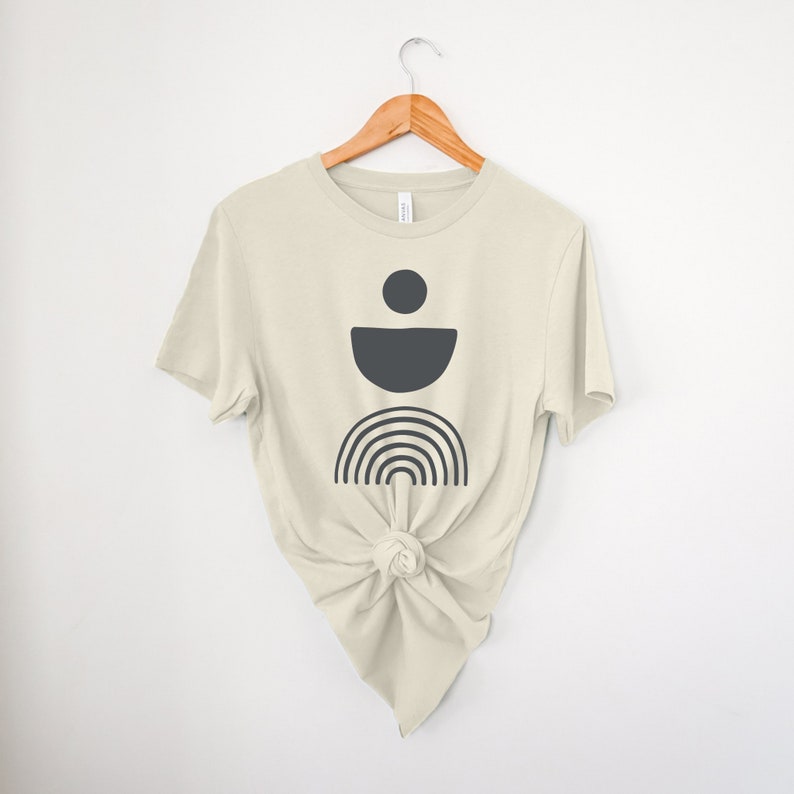May include: A cream-coloured t-shirt with a dark grey geometric design featuring a circle, a half-circle, and a rainbow. The t-shirt is knotted at the waist and hangs on a wooden hanger against a white background.