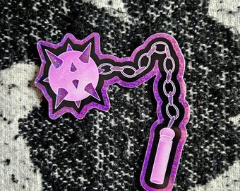May include: A purple and black glitter sticker of a spiked ball on a chain with a handle. The sticker is on a black and white patterned fabric.