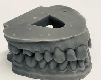 Puede incluir: Un modelo de mandíbula humana en 3D impreso en gris con dientes. El modelo está diseñado para uso dental y muestra la disposición de los dientes en la boca.