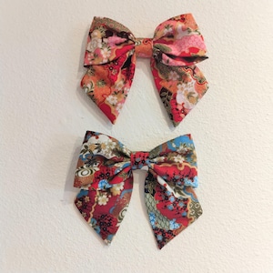 May include: Two fabric hair bows with a Japanese floral pattern. The bows are red with accents of pink, blue, and gold. The bows are tied in a classic bow shape and are likely handmade.