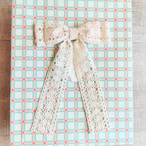 Könnte beinhalten: Eine Geschenkbox, verpackt in gemustertem Papier mit einer cremefarbenen Spitzen-Schleife. Das Papier hat ein sich wiederholendes Muster aus hellblauen Quadraten mit korallenfarbenen Kreisen. Die Schleife ist aus zarter, offen gewebter Spitze.