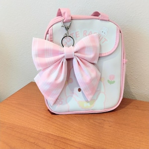 May include: A white and pink rectangular bag featuring a large pink and white plaid fabric bow attached to a metal clip. The bag has pink trim, a top handle, and a graphic print with the text "CUTE RABBIT" and a small flower illustration.