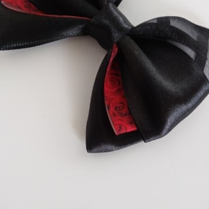 May include: A black satin bow with a red and black patterned ribbon underneath. The bow is tied with a loop and a knot.