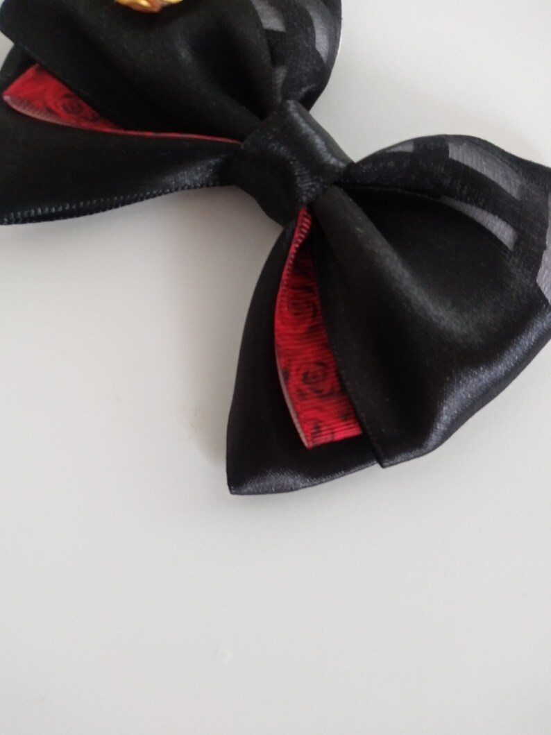 May include: A black satin bow with a red and black patterned ribbon underneath. The bow is tied with a loop and a knot.