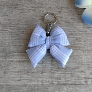 May include: A fabric bow keychain featuring a light blue and white plaid pattern. The bow is attached to a silver-toned metal split ring and a lobster claw clasp, providing a secure way to attach the accessory to bags, keys, or other items.