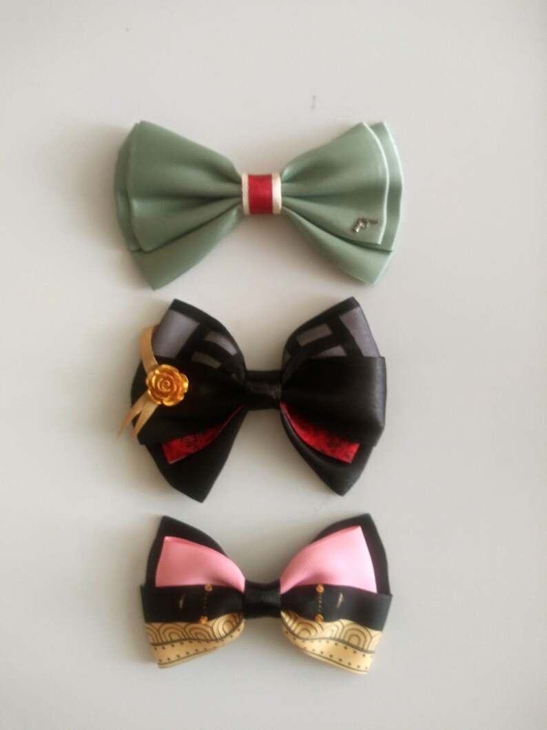 May include: Three different bow ties. The top bow tie is green with a red stripe. The middle bow tie is black with a red and gray pattern and a gold flower accent. The bottom bow tie is black with a pink and gold pattern.