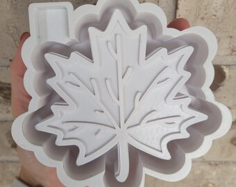 Puede incluir: Un cortador de galletas de plástico blanco en forma de hoja de arce. El cortador tiene un diseño de hoja detallado con venas visibles y un borde festoneado. El cortador se sostiene en una mano, con un reloj blanco visible en el fondo.