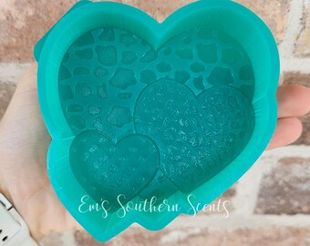 May include: A teal heart-shaped mold with three heart-shaped impressions. The largest heart has a leopard print pattern, while the smaller hearts have text. The mold is held in a hand. The text "Emi's Southern Scents" is visible.
