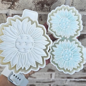 May include: A set of three white plastic sunflower-shaped cookie cutters. The cutters feature detailed petal designs and star accents in the center. The largest cutter measures approximately 10 cm in diameter.