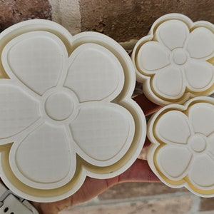 May include: Three white plastic flower-shaped cookie cutters of different sizes. The largest cutter has a double layer design.