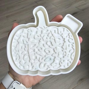 May include: A white plastic pumpkin-shaped cookie cutter with a detailed, textured design. The cutter has a stem and a rectangular tab on the side. Ideal for fall baking and Halloween treats.