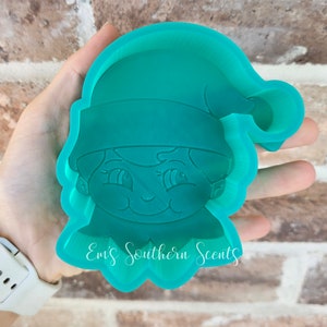 Puede incluir: Un molde de silicona verde turquesa de un elfo con un gorro de Papá Noel. El molde tiene la forma de la cabeza y los hombros de un elfo, con una gran sonrisa y ojos grandes. El molde es para hacer manualidades de resina.