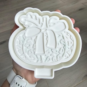 Op de afbeelding: Witte plastic pompoenvormige koekjesuitsteker met een strik- en bladerontwerp. De pompoen heeft een textuurpatroon. De uitsteker wordt in een hand gehouden.
