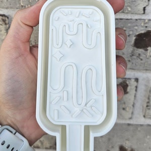 Może przedstawiać: Biała plastikowa forma w kształcie lodów na patyku z falistym wzorem i posypką. Forma jest przeznaczona do robienia domowych lodów na patyku.