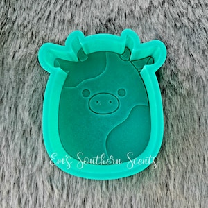 Op de afbeelding: Een groene siliconen mal in de vorm van een cartoon-koe met zwarte vlekken. De mal ligt op een grijs, pluche oppervlak.