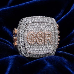 May include: A large, square-shaped ring featuring a dense pavé setting of clear, sparkling stones covering the entire surface. The letters GSR are prominently displayed in the centre, outlined in rose gold and filled with small stones.