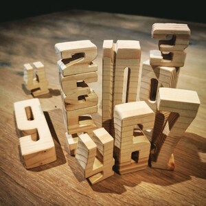 May include: Wooden number blocks in various sizes and shapes, arranged on a wooden surface. The numbers 1, 2, 3, 4, 6, 7, and 9 are visible. The blocks are made of light-colored wood and have a natural finish.
